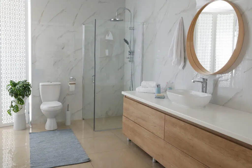Modern bathroom with marble walls, glass shower enclosure, wooden vanity, and round mirror.